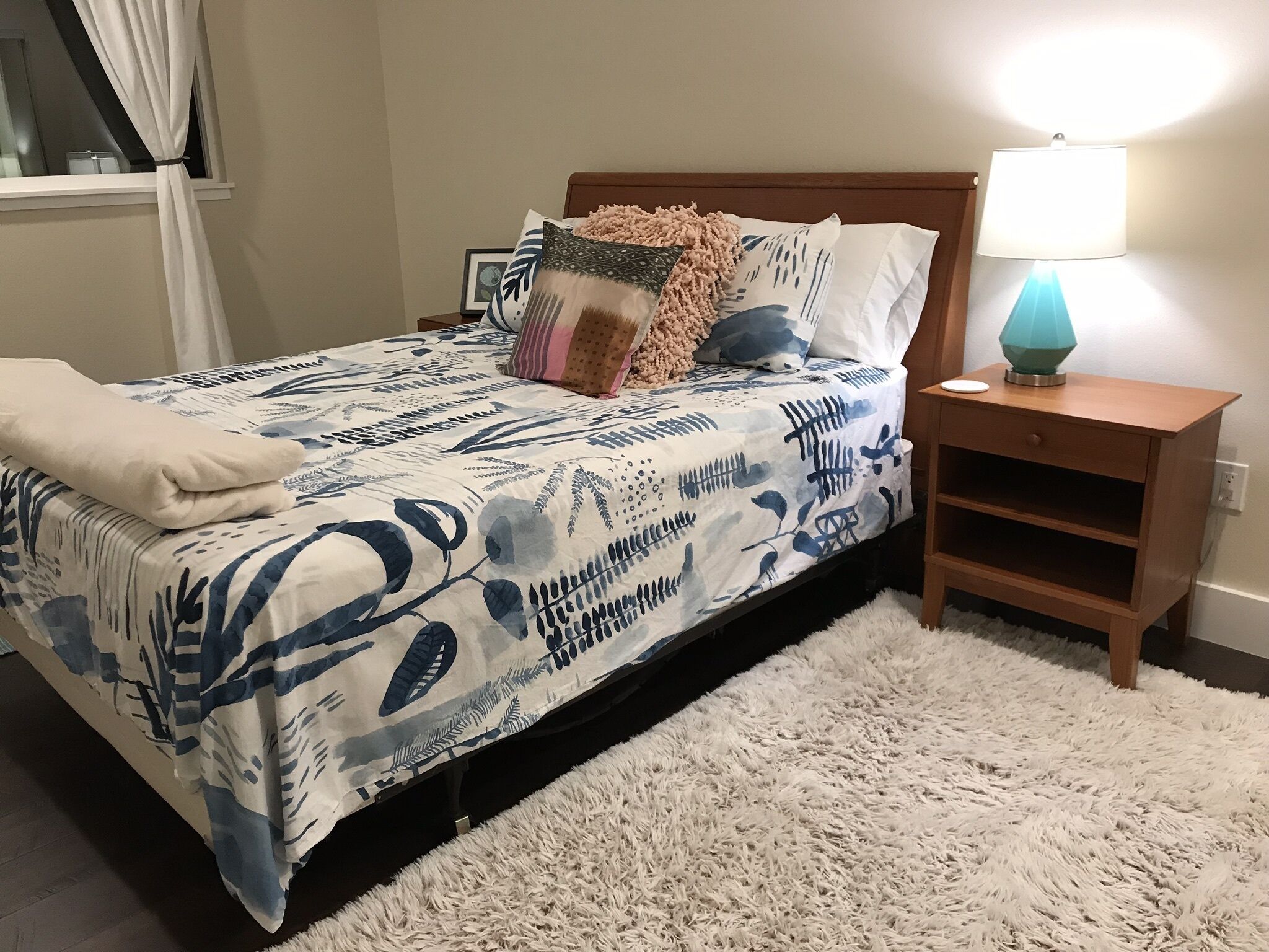 A neatly made bed with a blue and white leaf-patterned bedspread, a fluffy white rug, and a bedside table with a lamp.