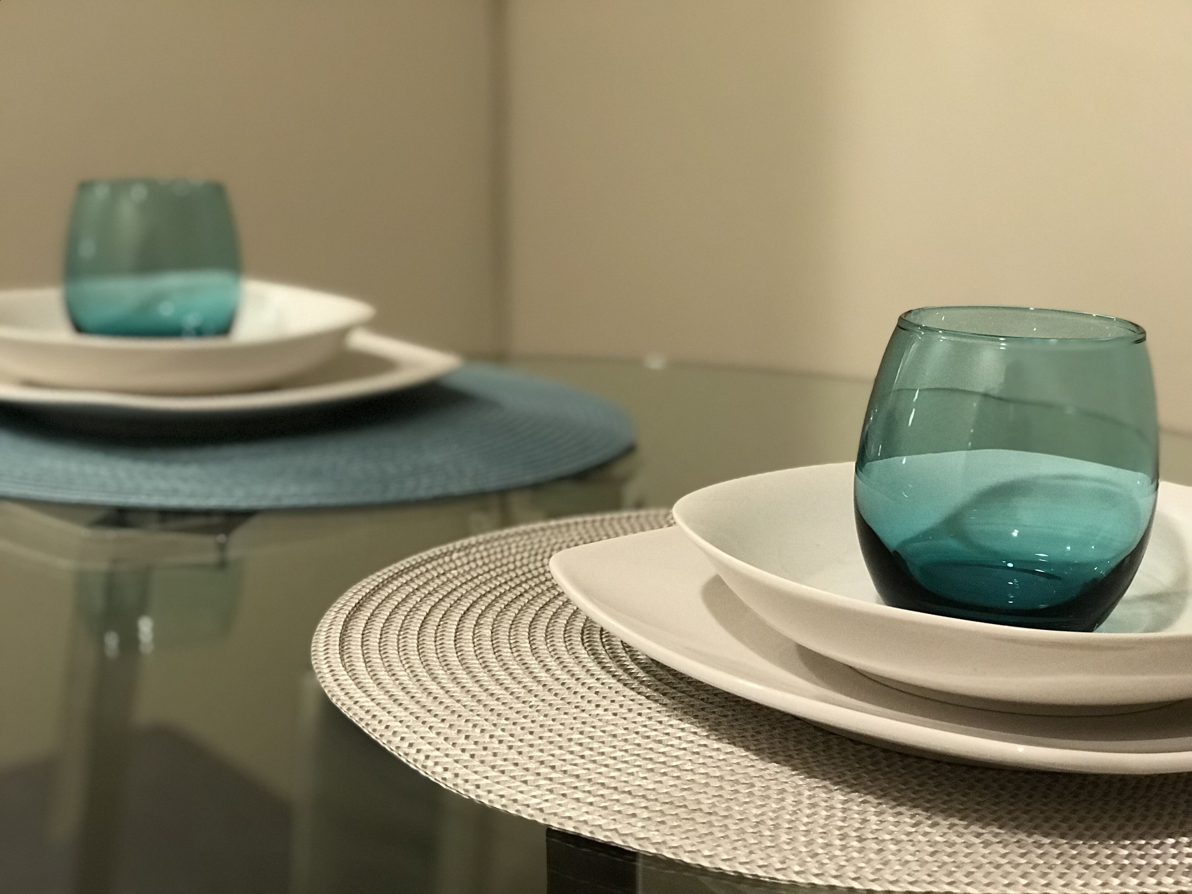 A dining table with a glass top, white plates, and a woven placemat.