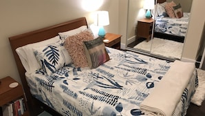 A neatly made bed with a patterned bedspread, a bedside lamp, a nightstand with a book, and a mirror reflecting the room. - DaLight - A beautiful home in a quiet neighborhood (Kailua)