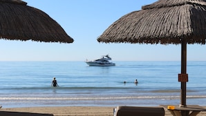 Ein Strand mit Reetdachsonnenstühlen, einem Rettungsschwimmturm und einer Yacht im Wasser.