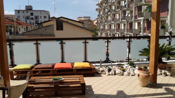 A terrace with wooden benches and cushions, a tiled floor, and a view of a water body and buildings in the background. - Superb Mansarda ... (Caserta)