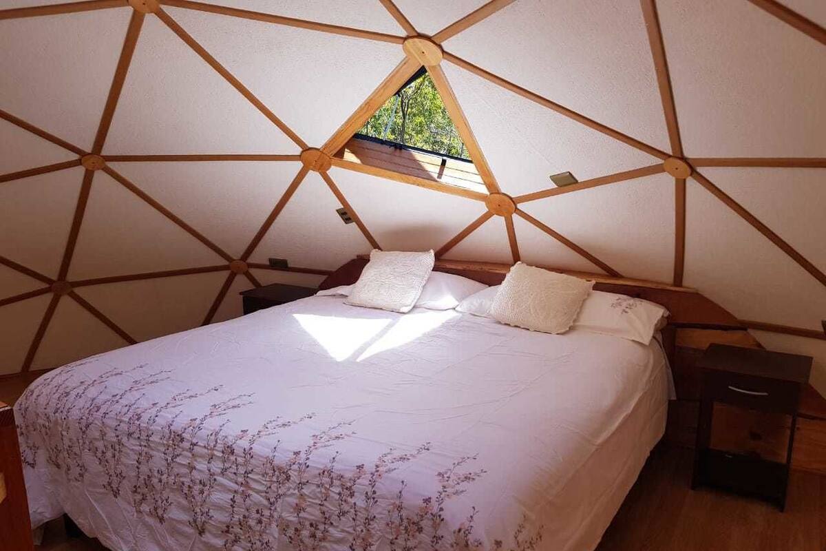 A bedroom with a geometric wooden ceiling structure, a bed with floral bedding, and a small window.