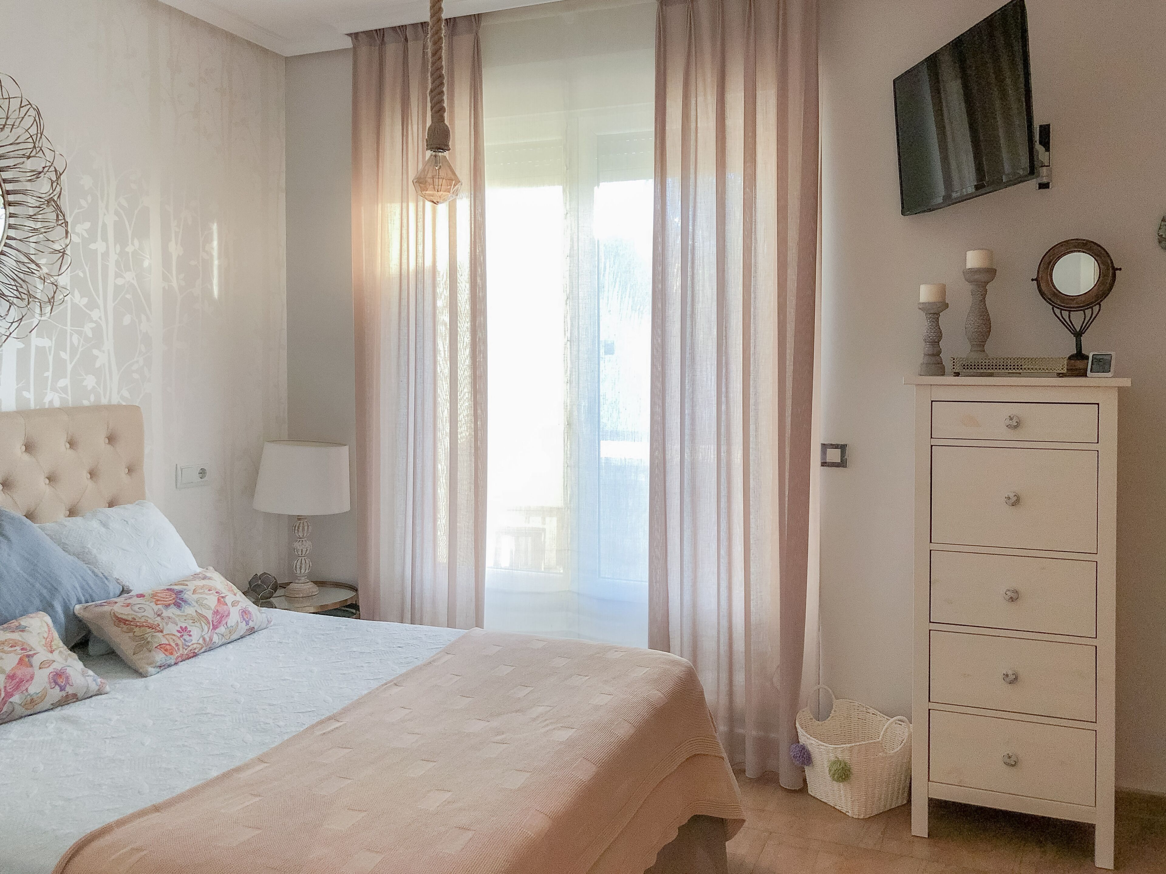 A bedroom with a bed, a nightstand, a dresser, and a window with sheer curtains.