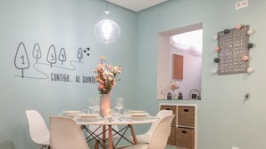 Un coin repas avec une table ronde dressée pour quatre personnes, des chaises blanches, un vase de fleurs et un mur orné d’une affiche présentant les règles familiales.