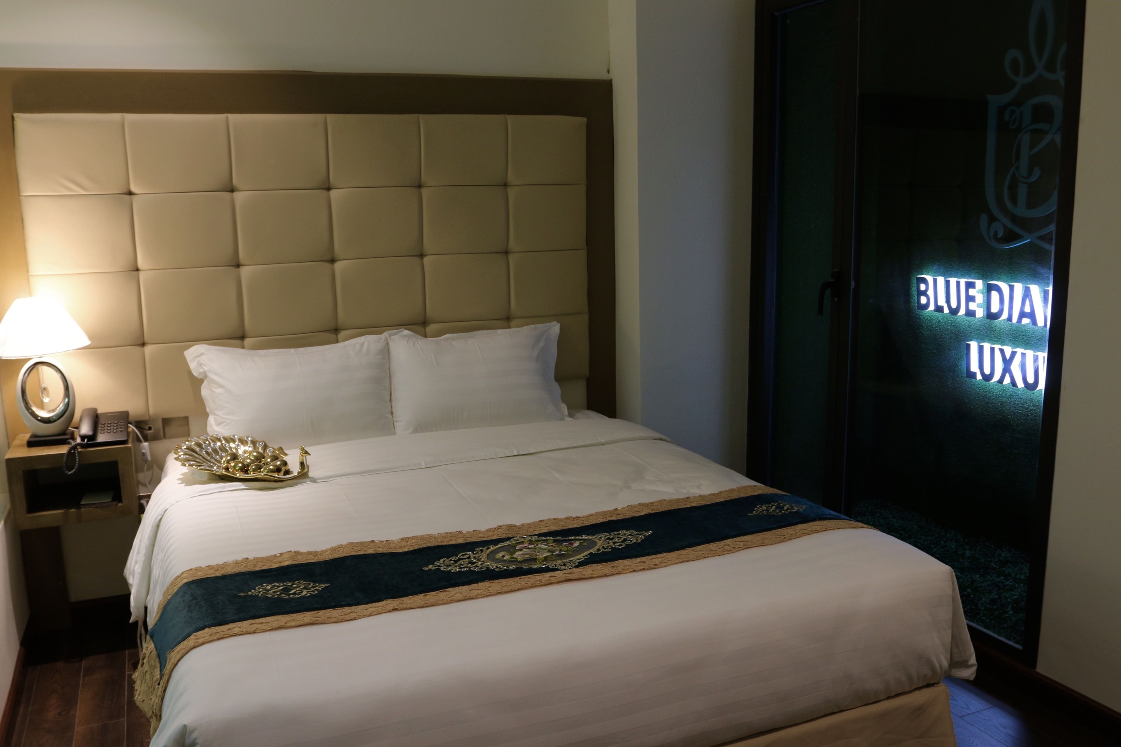A hotel room with a large bed, a bedside table with a lamp, and a door with a sign that reads "BLUE DIAMOND LUXURY".