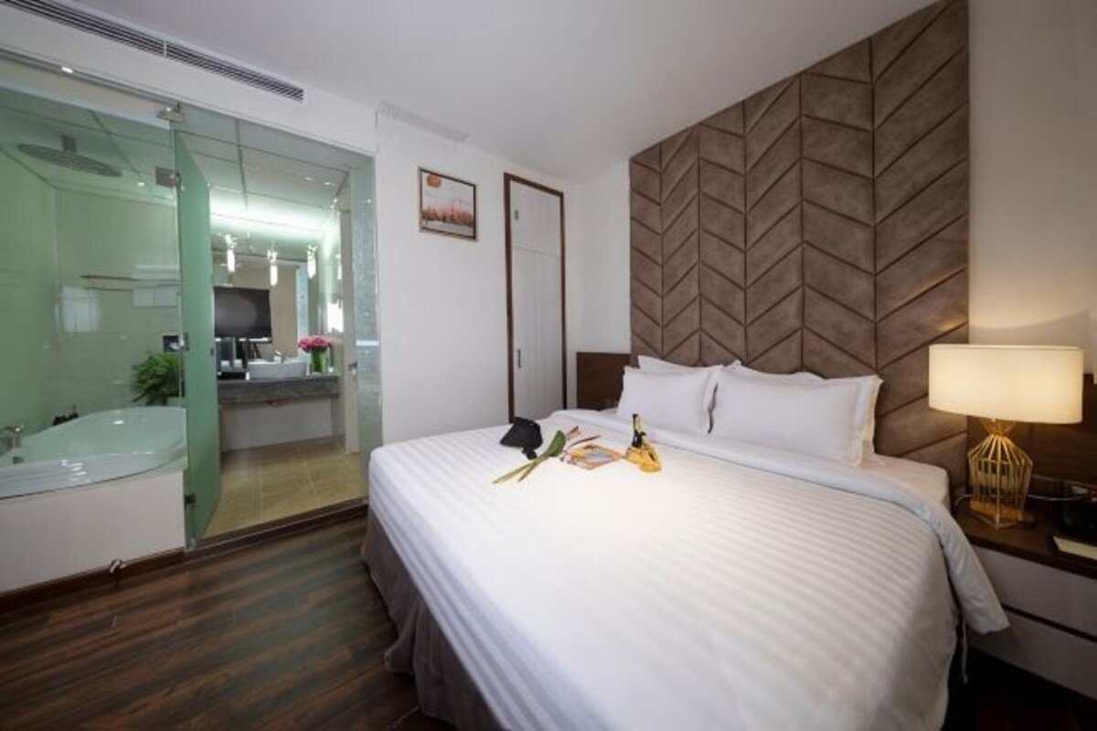 A hotel room with a bed, bedside table, lamp, and a view of a bathroom with a mirror and sink.