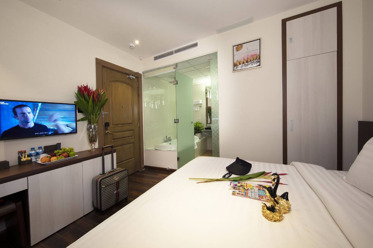 Een hotelkamer met een bed, een televisie, een bureau met een bloevas, een koffer en een badkamer die door een glazen deur zichtbaar is.