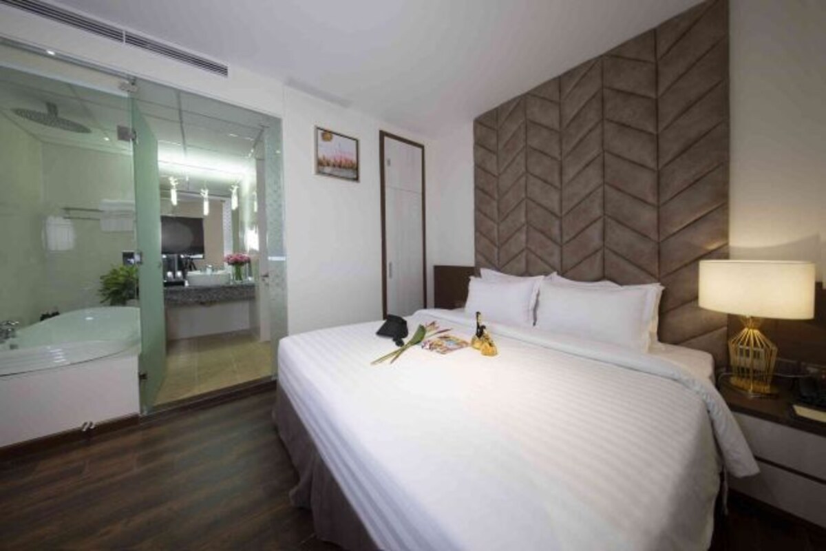 A hotel room with a bed, a bedside lamp, a mirror, and a bathroom visible through a glass door.
