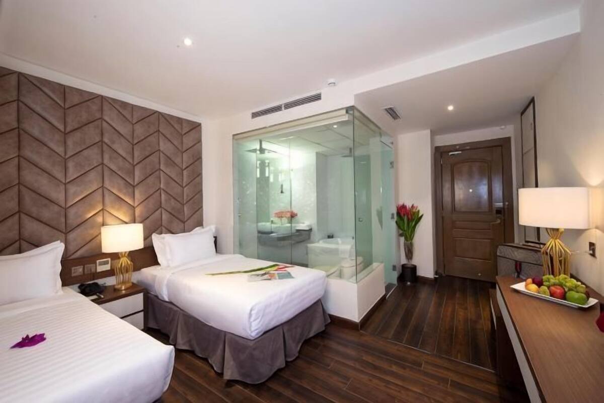 Een hotelkamer met twee bedden, een douchecabine met glazen wanden, een houten vloer en een bureau met lamp.