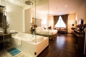 Una habitación de hotel moderna con una bañera grande, un dormitorio independiente con cama, una zona de estar con sofá y un pequeño escritorio con lámpara.