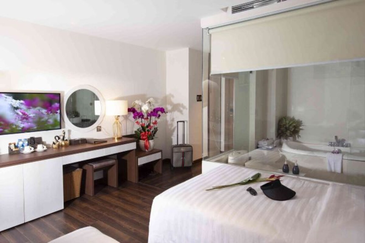 A hotel room with a bed, a desk with a mirror, a TV, a bathroom with a bathtub, and a large window.