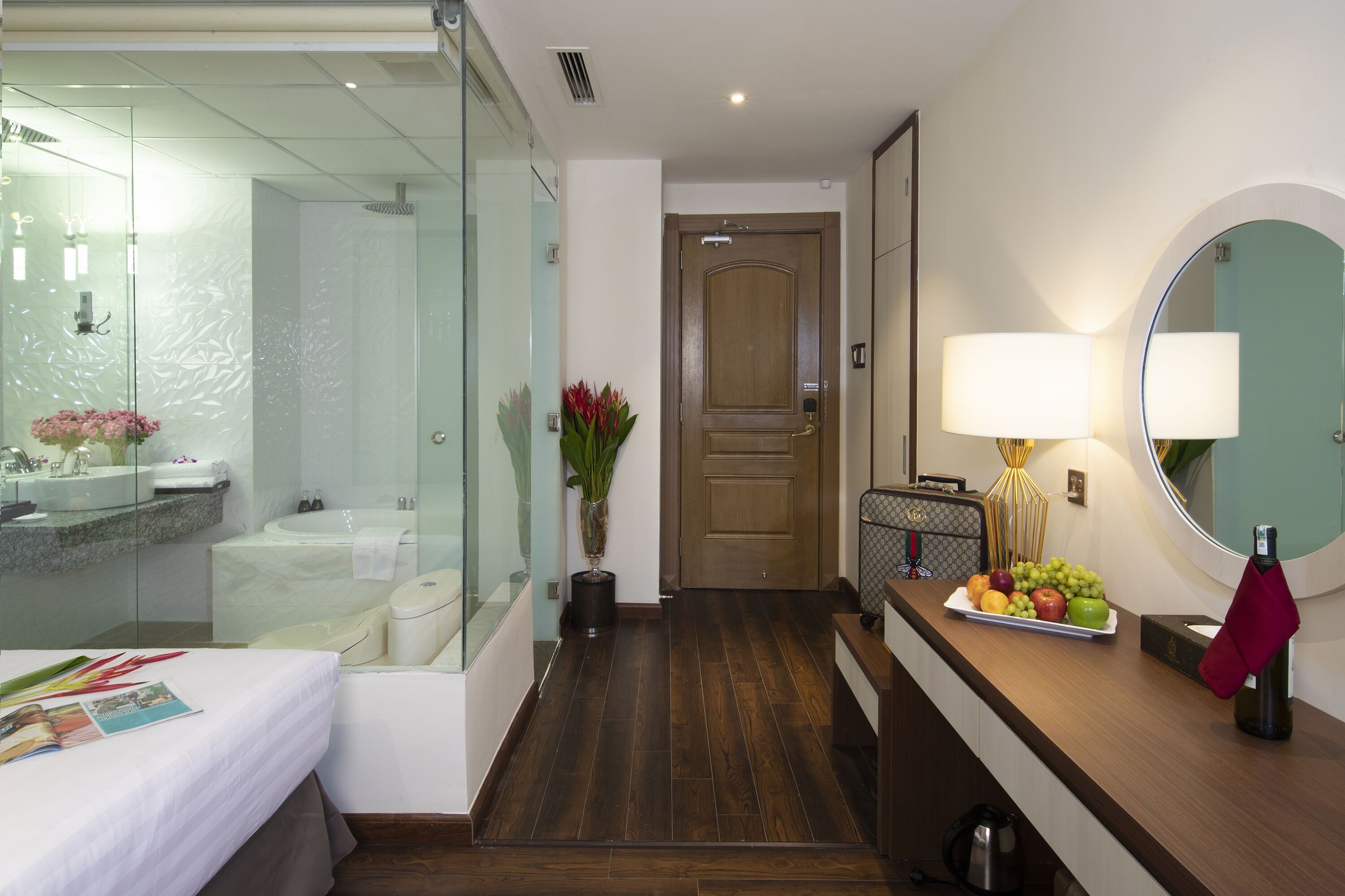 Une chambre d’hôtel avec une douche en verre, une porte en bois, une table de chevet avec une lampe et une assiette de fruits posée sur le comptoir.