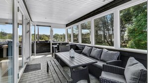 Eine Terrasse im Freien mit einem Holztisch und Korbmöbeln, große Fenster mit Blick auf Bäume und einen klaren Himmel.
