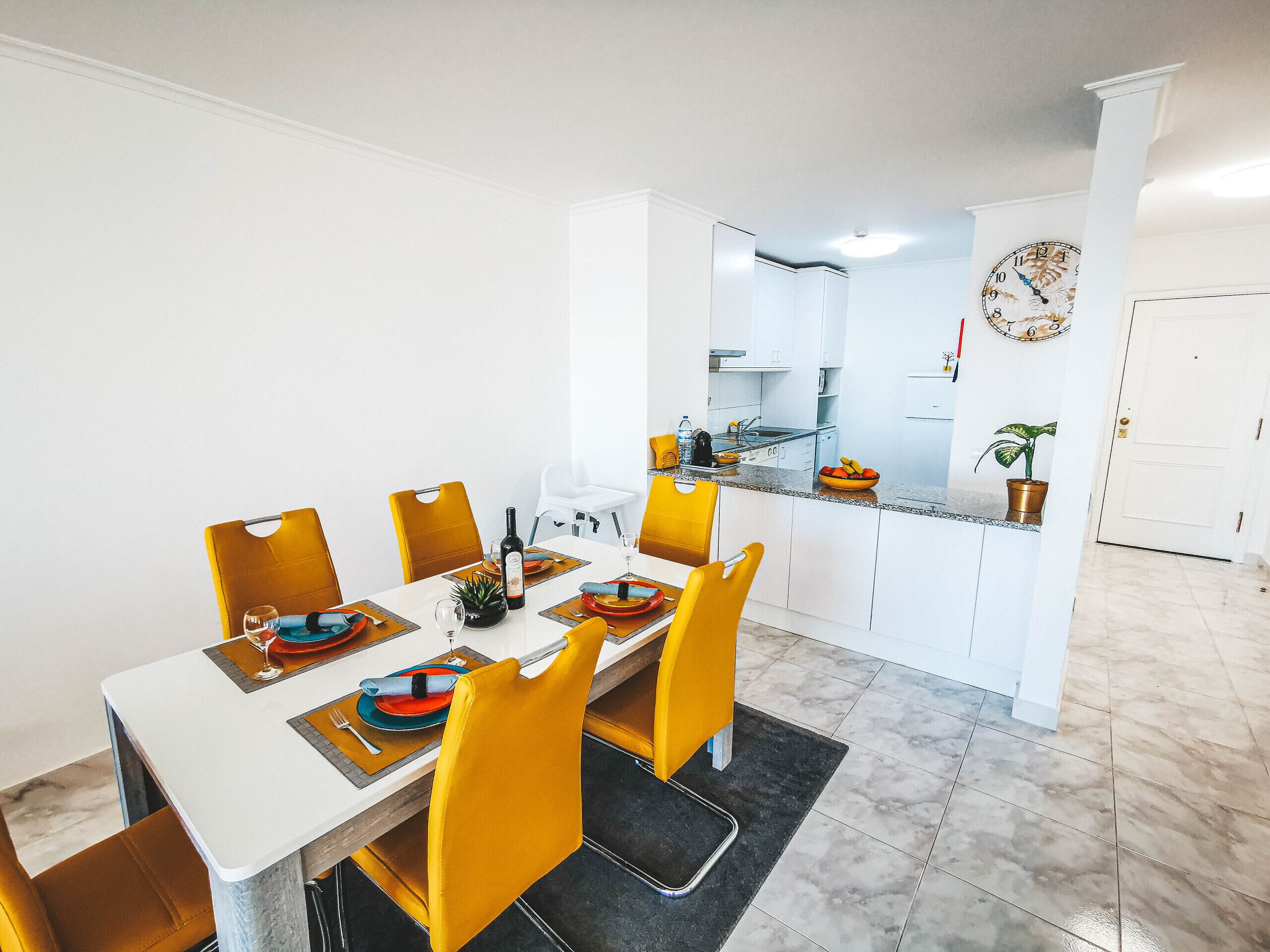 A dining area with a white table, yellow chairs, and a kitchen in the background.
