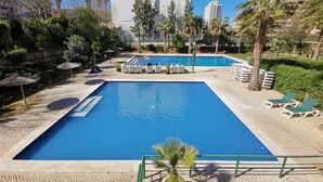 A swimming pool with lounge chairs and umbrellas, surrounded by greenery and tall buildings.