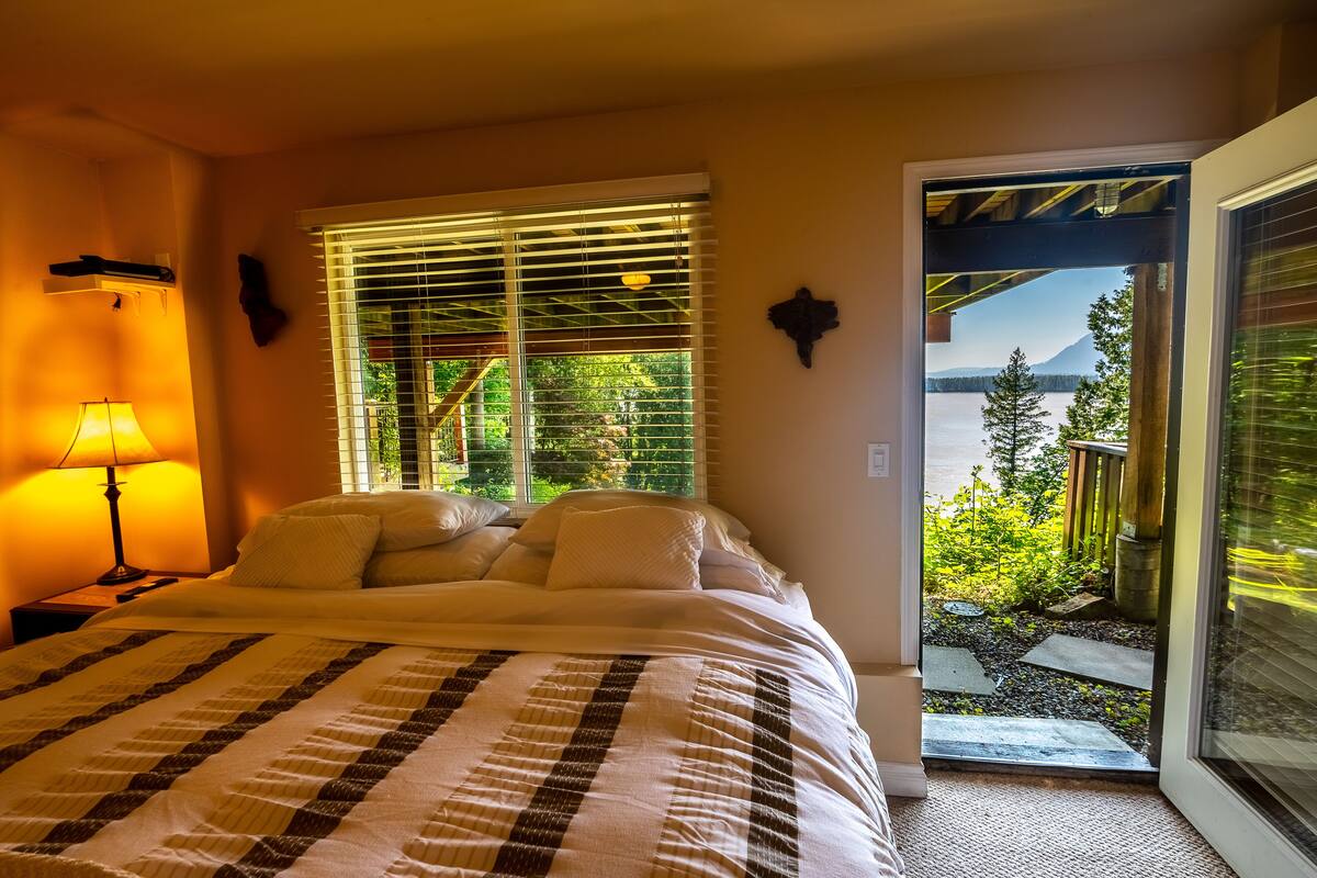 Un dormitorio con cama, ventana con persianas, mesita de noche con lámpara y vistas a un jardín y montañas a través de la ventana.