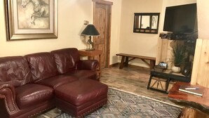 A living room with a brown leather sofa, a patterned rug, a wooden bench, a small table with a lamp, a framed horse painting, and a television on a wooden stand. - The Corral 2  room apartment next to The Yellowstone Rive (Gardiner)