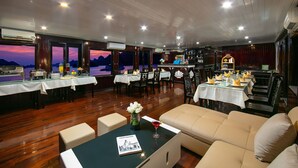 A restaurant with wooden floors, tables set for dining, and a view of the mountains at sunset. - Halong Fantasea Cruise (Tuan Chau)