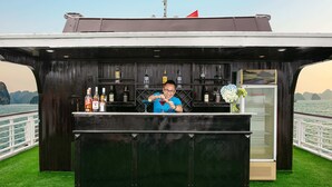 A bar area with a bartender, shelves of liquor bottles, and a menu stand. - Halong Fantasea Cruise (Tuan Chau)