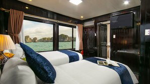 A double bed with white and blue bedding, a nightstand with a lamp, and a view of the sea through large windows. - Halong Fantasea Cruise (Tuan Chau)