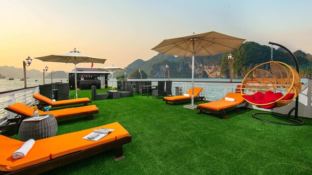 A rooftop lounge area with artificial grass, sun loungers, a bar, and a hanging chair overlooking a body of water and mountains. - Halong Fantasea Cruise (Tuan Chau)