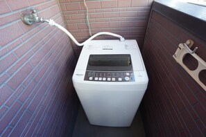 Een compacte, witte Hisense wasmachine in een kleine, afgesloten ruimte met een bakstenen muur en een metalen deur.