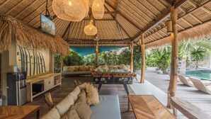 Un área de estar al aire libre cubierta con mobiliario de madera, una barra con refrigerador y una piscina al fondo.