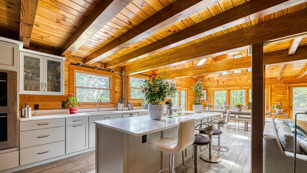 A modern kitchen with wooden beams, a large island, bar stools, and a dining area with a sofa and table. - Luxurious & Enchanting Log House in Heart of Hudson Valley/Catskills, Upstate NY (Saugerties)