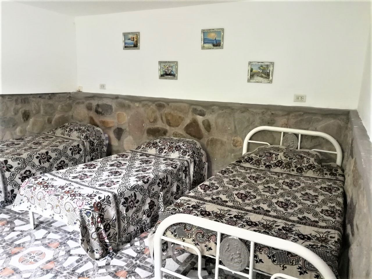 A room with three beds, each with patterned bedding, a stone wall, and small framed pictures on the wall.