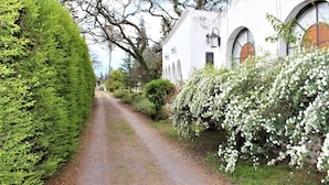 A narrow dirt path flanked by lush greenery and white flowering shrubs, leading to a white building with arched windows and a lantern. - Quinta Valvanera (Salta)