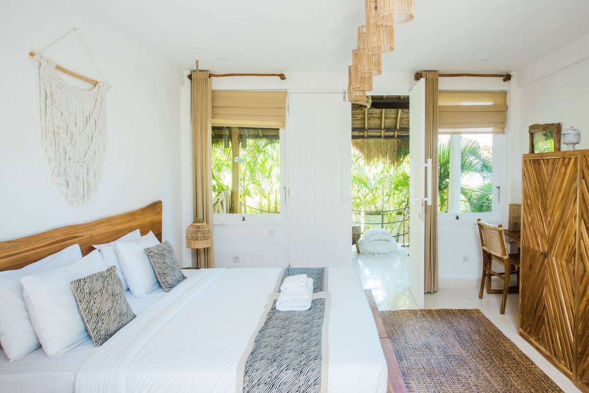 A bedroom with a bed, a wooden headboard, a macrame wall hanging, a window with curtains, a wooden wardrobe, and a view of greenery outside.