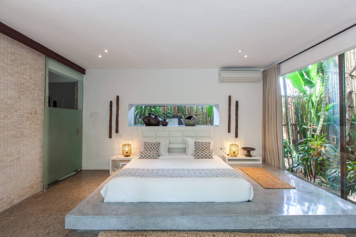 A modern bedroom with a large bed, a view of greenery through the window, and a minimalist design.