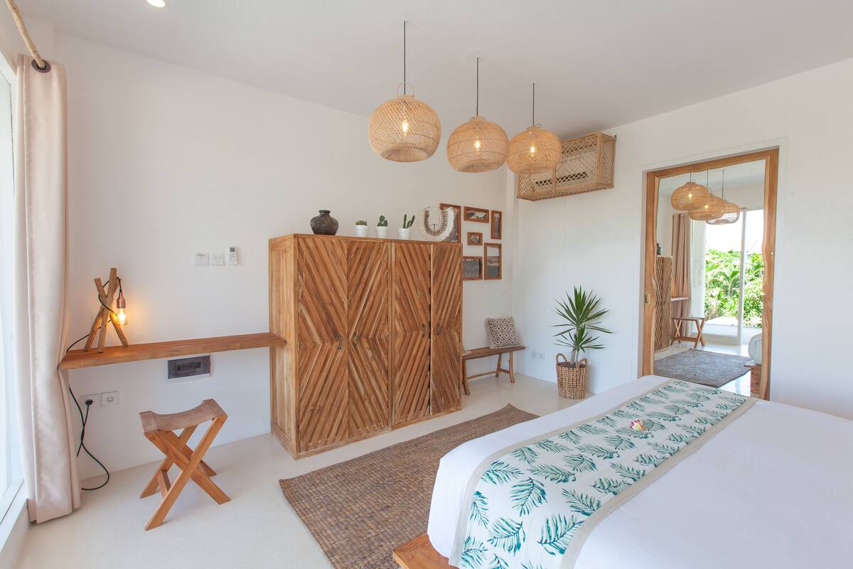 A bedroom with a bed, wooden wardrobe, bedside table, hanging lights, and a view to the outside.