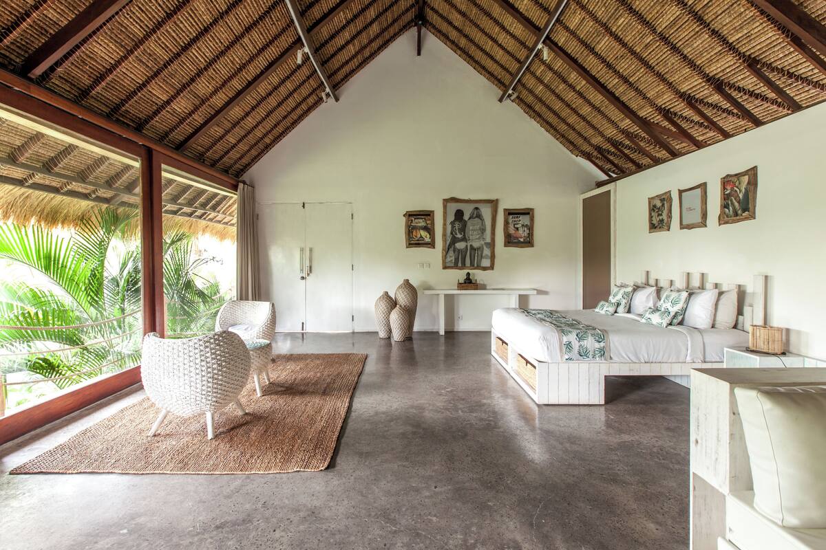 A spacious room with a high ceiling, a large window with a view of palm trees, a bed with white bedding, a white sofa, and a small table.