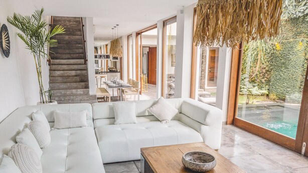 A modern living room with a large white sectional sofa, a wooden coffee table, and a view of an outdoor swimming pool. - AB Villa & Apartments (Seminyak)