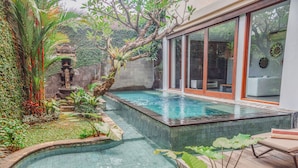 A swimming pool with a wooden deck and a small hot tub, surrounded by lush greenery and a stone wall. - AB Villa & Apartments (Seminyak)