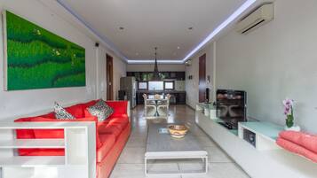 Una sala moderna con un sofá rojo, una mesa de centro y un televisor. Se ve una zona de comedor con una mesa y sillas al fondo.