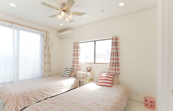 A bedroom with two beds, a ceiling fan, a window with curtains, and a small bedside table.