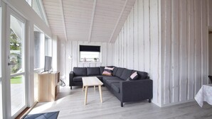 Una sala de estar moderna con un sofá seccional gris, una mesa de centro de madera y un televisor sobre un mueble de madera. Amplias ventanas ofrecen luz natural y vistas al exterior.