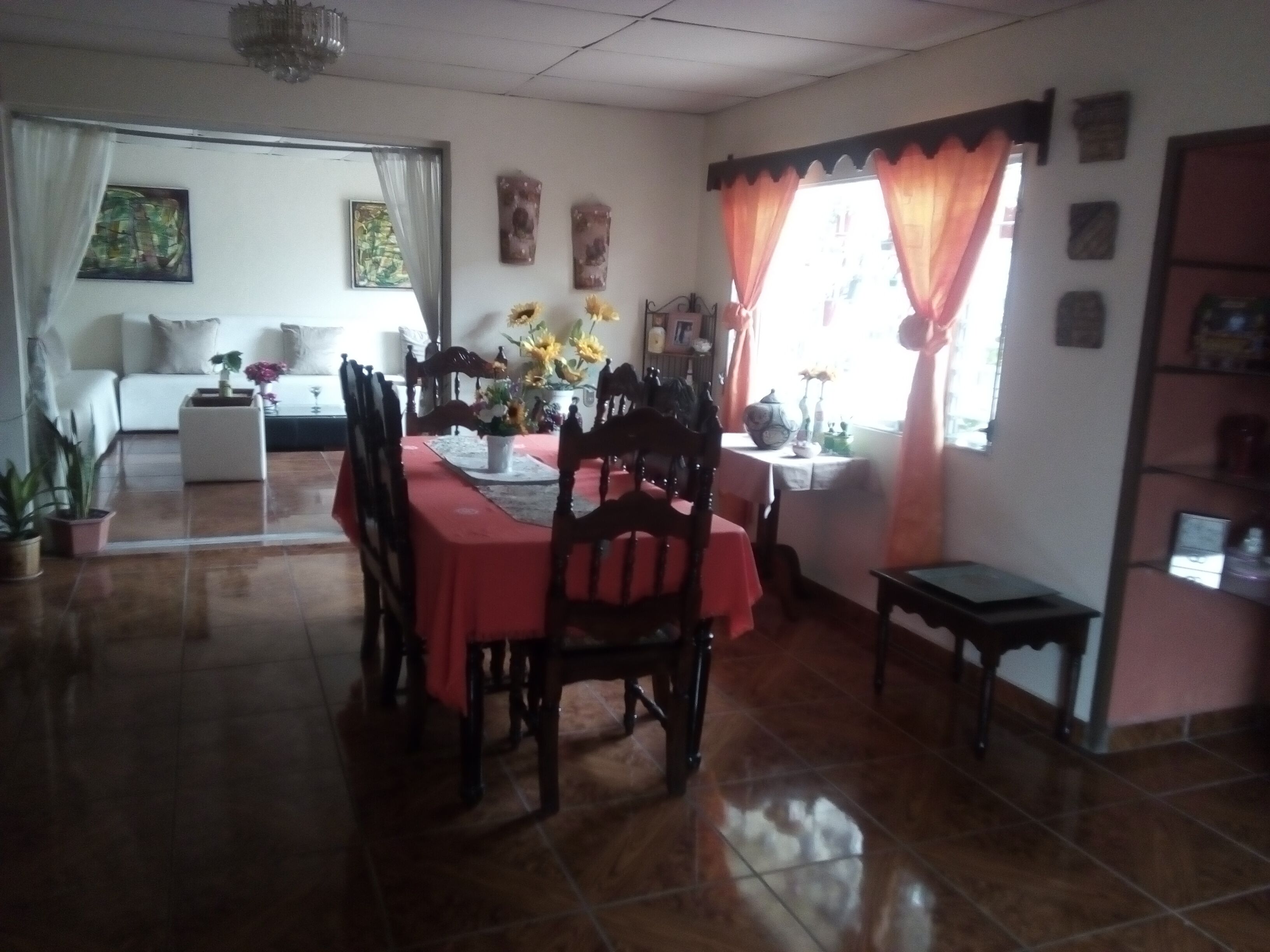 A dining area with a red tablecloth, wooden chairs, and a vase of flowers. There is a sofa and a window with curtains in the background.