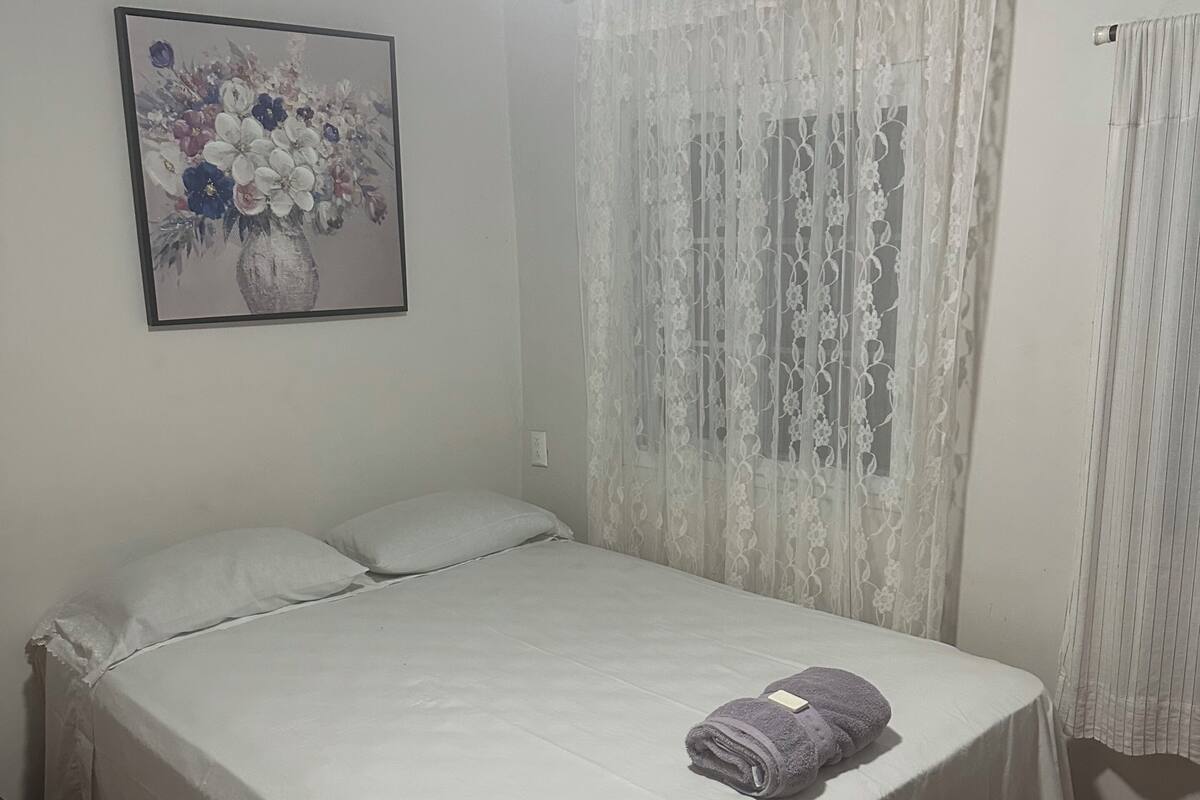 Una cama individual con una colcha blanca, una toalla enrollada y un cuadro floral en la pared.