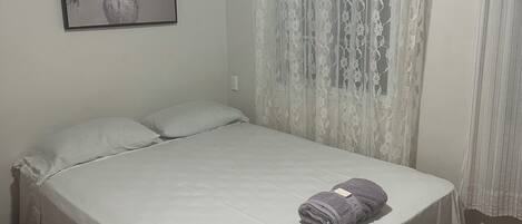 Una cama individual con una colcha blanca, una toalla enrollada y un cuadro floral en la pared.
