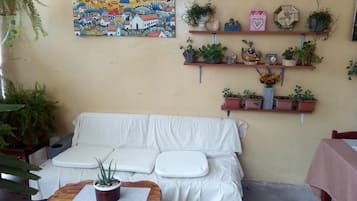 A cozy room with a white sofa, a small wooden table with a plant, and a wall decorated with a colorful painting and shelves with potted plants.