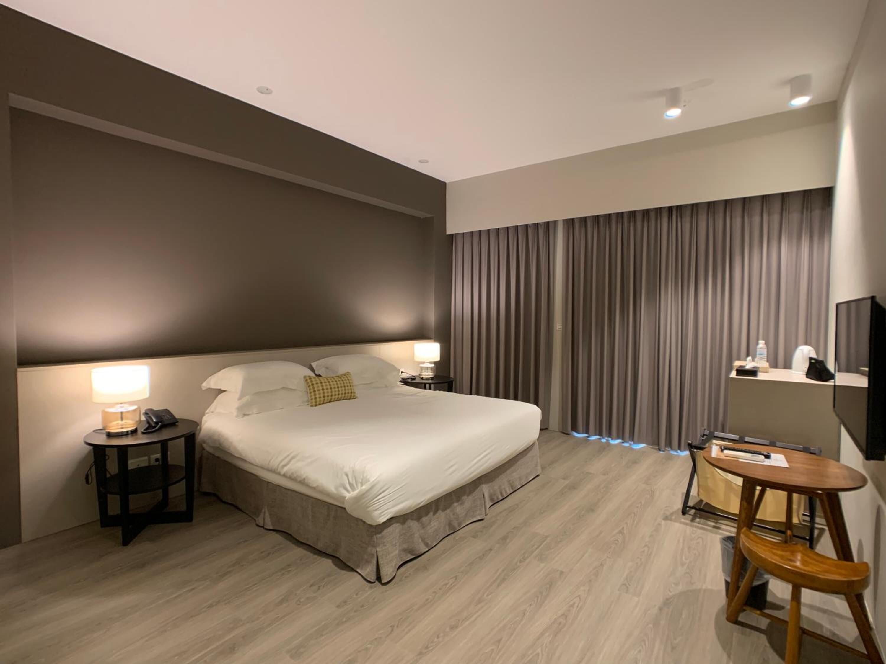 Una habitación de hotel moderna con una cama grande, mesitas de noche con lámparas, un escritorio con silla y un ventanal con cortinas.