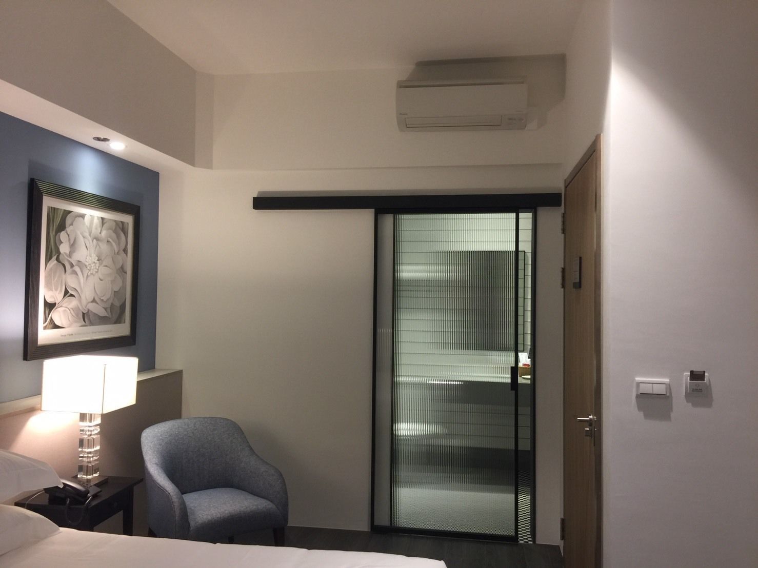Een hotelkamer met een bed, een stoel, een bureau met een lamp, een ingelijst schilderij aan de muur, en een schuifdeur met ruitfolie.