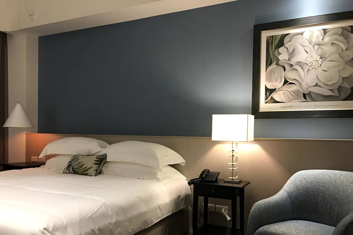 A hotel room with a bed, a nightstand with a lamp, a chair, and a framed artwork on the wall.