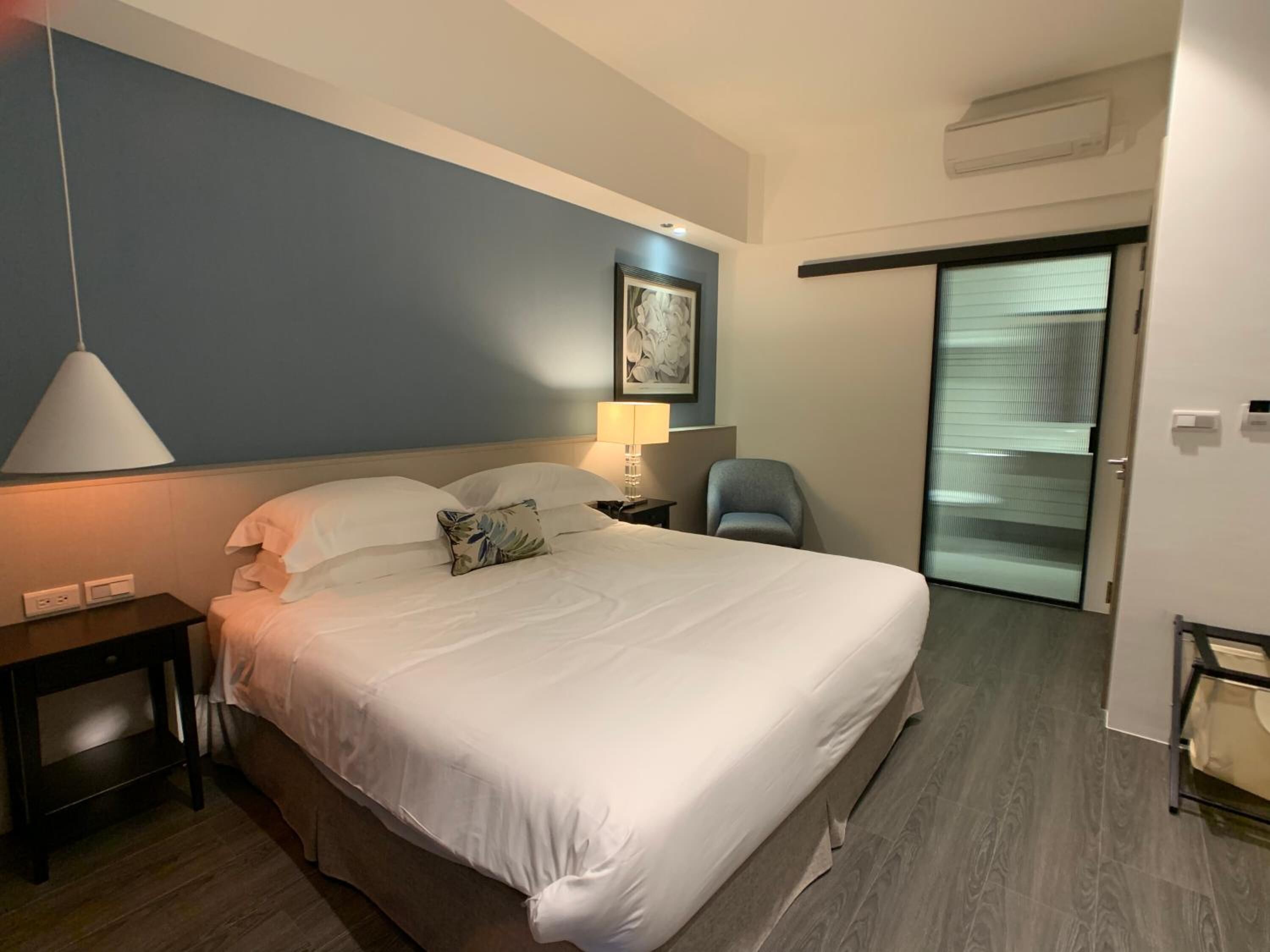 Un cuarto de hotel con una cama grande, una mesita de noche, una lámpara, una silla y un cuadro en la pared.