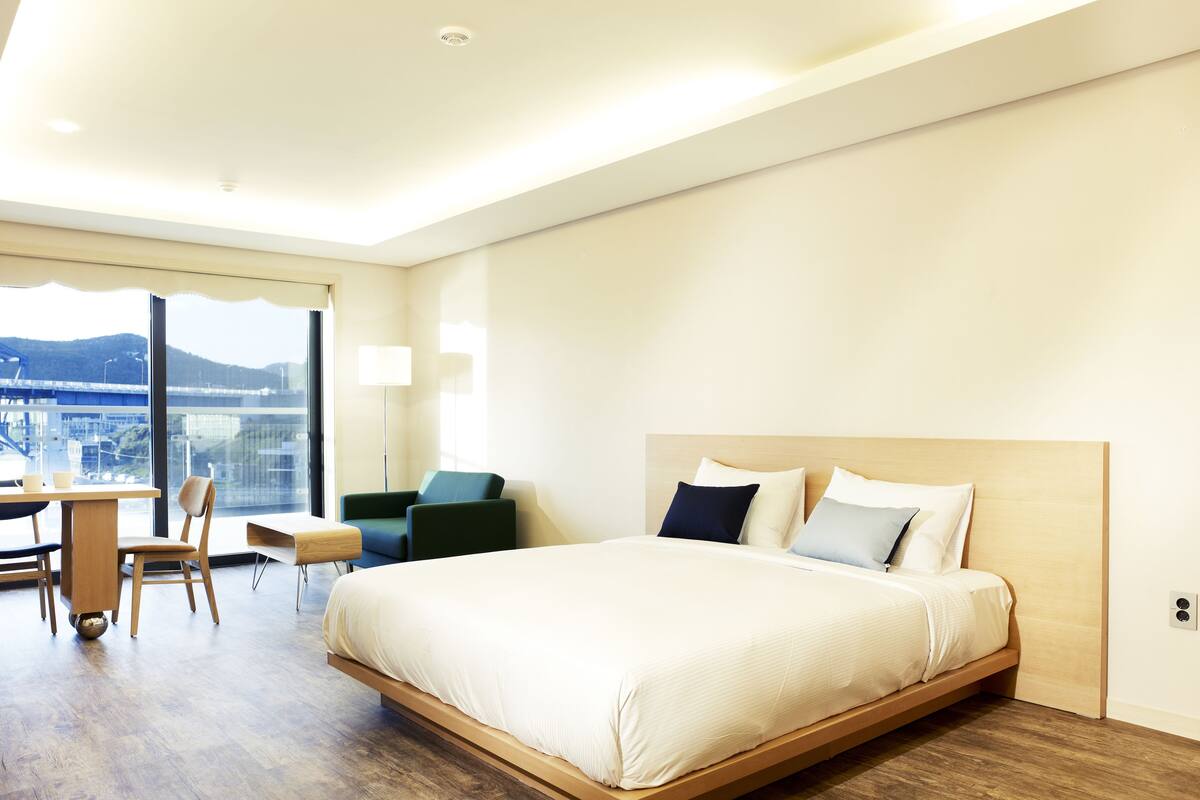 A modern hotel room with a large bed, a desk with a chair, and a view of a bridge and cityscape through large windows.
