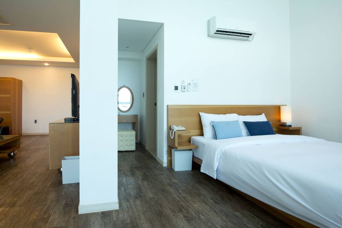 A hotel room with a bed, bedside table, lamp, and an air conditioning unit.
