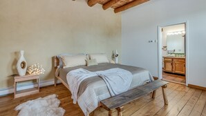 Un dormitori amb sostre de fusta, un llit amb llençol gris, una panxa, un feser de pell blanca i una tauleta de nit amb un ram i llums.